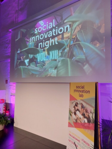 Eine große weiße Wand, auf die ein Beamer den Schriftzug Social Innovation Night Vol.XIII  wirft. Davor steht ein Aussteller des Social Innovation Lab.