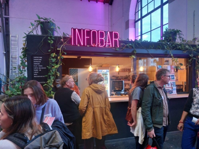 Rosafarbener Leuchtschriftzug "Infobar" auf einem Häuschen. Davor stehen Menschen, die sich dort etwas zu Trinken kaufen.