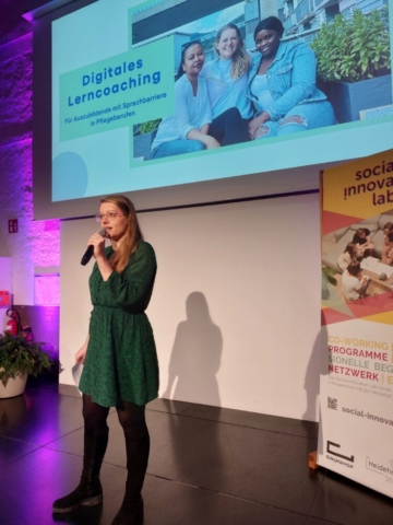 Sarah Huber vom Social Startup Learncoaching steht auf der Bühne und spricht in ein Mikrofon. Sie stellt ihr Startup vor.