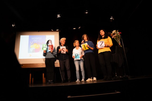 Fünf Menschen, davon drei Frauen und ein Mann, stehen auf der Bühne. Jede*r hält einen Buchstaben hoch, die zusammen das Wort "HOSPOZ" ergeben.