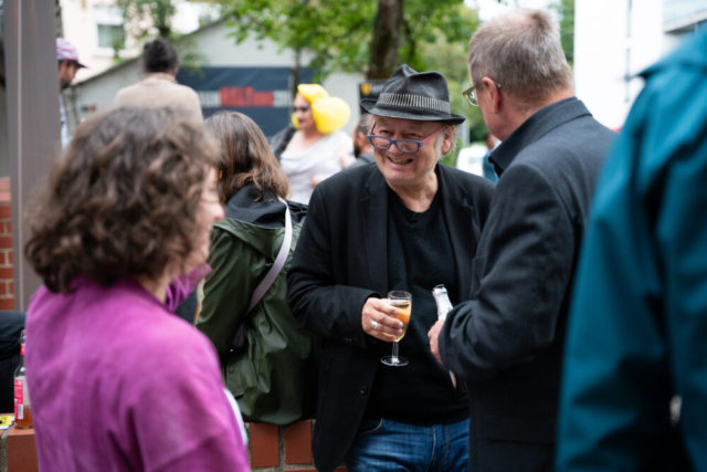 Ein Mann mit Brille und Hut hat ein Sektglas in der Hand und lacht zwei anderen Menschen zu