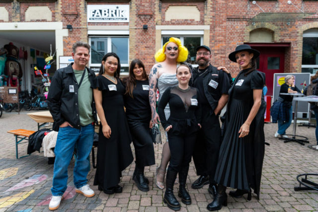 Die bunte Gruppe des Projekts "Gesichter gegen RechtS". Sieben Menschen, davon sechs schick in Schwarz und eine Drag-Queen mit gelber Marylin Perücke und silbernem Glitzerkleid.