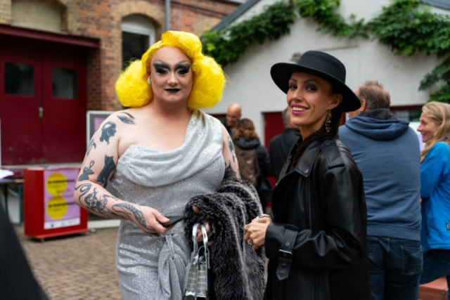 Drag Queen Data Tip mit gelber Perücke, auffälligem Make-up und silbernem Kleid. Daneben eine Frau in schwarzem Ledermantel und mit schwarzem Hut. Beide lächeln in die Kamera